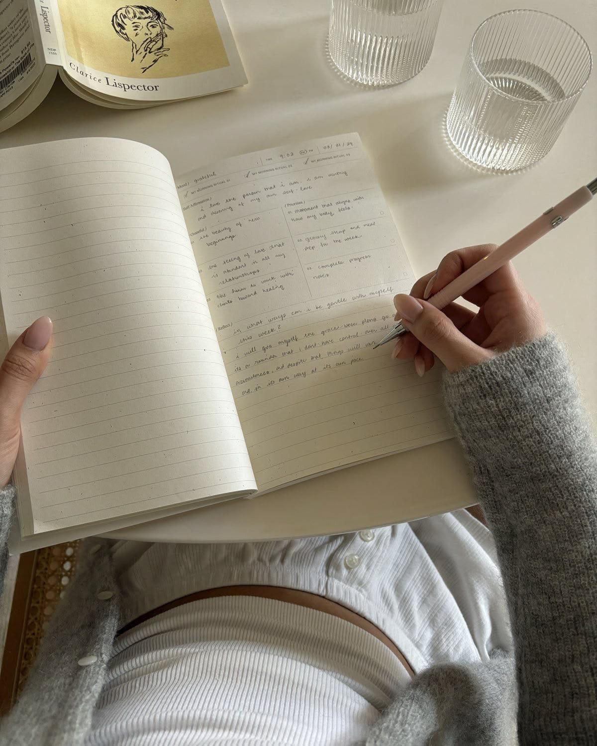Hænder, der skriver i en notesbog ved siden af en bog og vandglas
